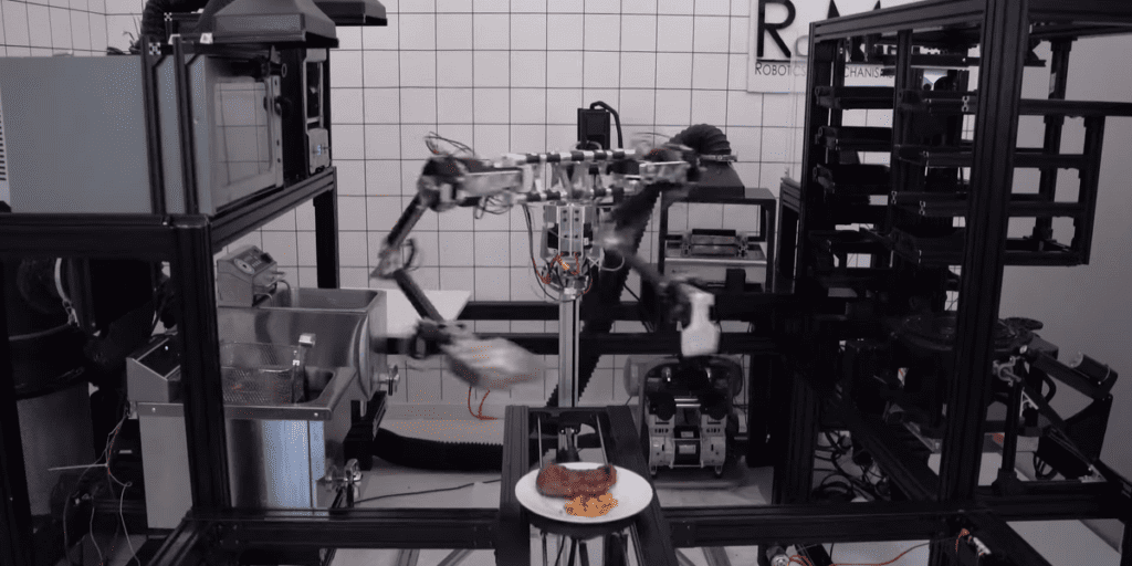 A Still From A Video Shows A Metal Robot With Arms Surrounded By Kitchen Equipment.png